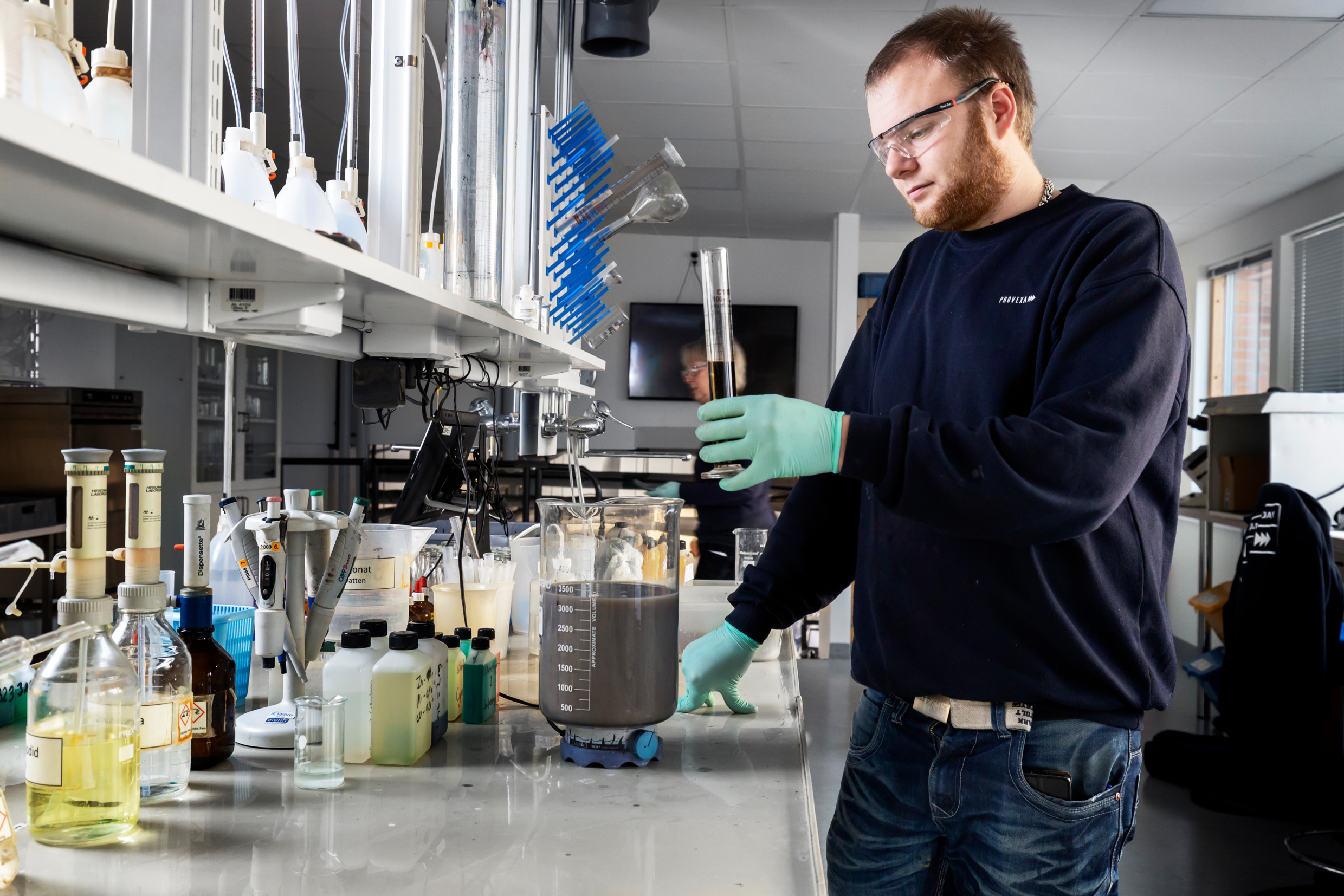 A man is holding a test tube with some liquid. The countertop in front of him is filled with bottles and containers with chemicals.